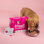 Dog eating from a bowl next to a box of Proudi dog food on a pink background