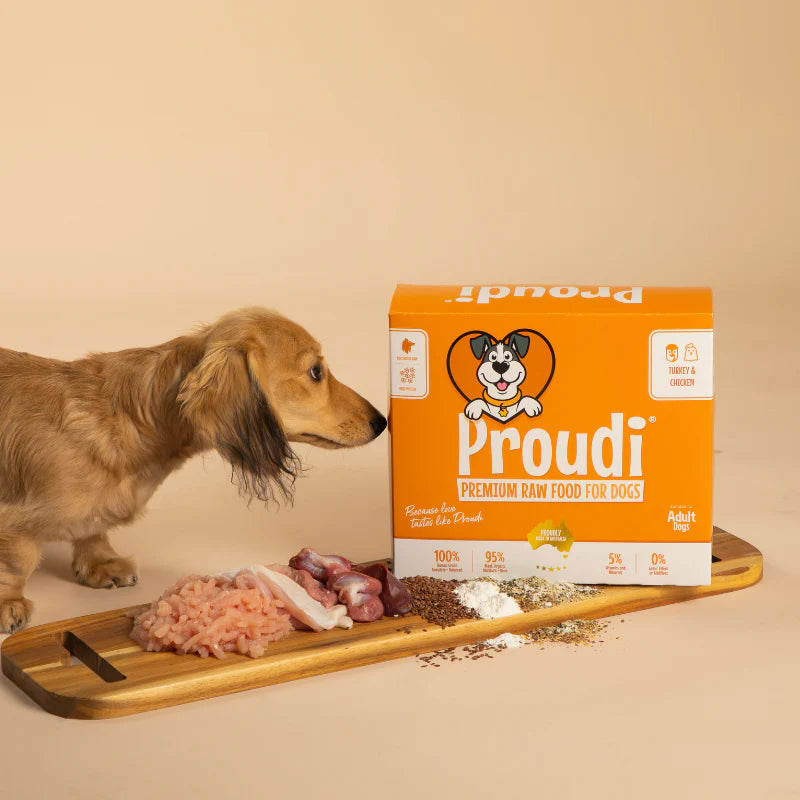 Proudi dog food box with a dachshund and raw dog food ingredients on a wooden board.